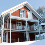 Ein modernes, zweistöckiges Haus mit einem steilen Dach ist mit Schnee bedeckt. Das Haus verfügt über große Fenster und einen umlaufenden Balkon. An der Seite ist ein Auto geparkt und im Hintergrund sind schneebedeckte Bäume auf einem Hügel an einem hellen, sonnigen Tag zu sehen.