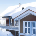 Ein zweistöckiges Haus mit großen Fenstern ist mit einer dicken Schneeschicht bedeckt, was auf einen kalten Wintertag hindeutet. Das Haus verfügt über einen umlaufenden Balkon und liegt vor einer Kulisse aus schneebedeckten Hügeln und einem klaren, blauen Himmel.