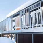 Ein modernes Gebäude mit großen Fenstern und einem Metallgeländer ist mit Schnee bedeckt. Das Dach und der Boden sind weiß bedeckt, was die Winterszene betont. Der Himmel ist klar und blau und im Hintergrund ist ein Teil eines anderen Gebäudes zu sehen.