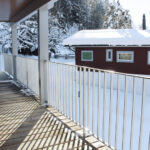Eine Holzveranda mit einem weissen Metallgeländer ist mit einer dünnen Schneeschicht bedeckt. Hinter dem Geländer sind ein schneebedeckter Hof und ein rotes Haus mit weissen Verzierungen zu sehen. Im Hintergrund sind schneebedeckte Bäume unter einem bewölkten Himmel zu sehen.