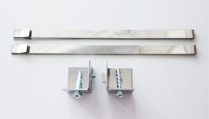 Zwei Metallbänder mit Aufschriften und zwei L-förmige Metallklammern mit Schrauben und Muttern sind auf einer weißen Fläche angeordnet. Auf einem Band steht „Delta“ mit einer Website-URL. Die Schrauben sind durch die Klammern gefädelt und bereit zur Montage.