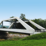 Eine moderne weisse Betonbrücke mit drei grossen geometrischen Stützen überspannt einen kleinen Fluss. Grünes Gras und üppige Bäume umgeben das Gebiet, und auf der linken Seite des Bildes ist ein Auto auf der Strasse zu sehen. Der Himmel ist bedeckt, was der Szene einen gedämpften Ton verleiht.