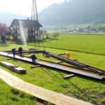 Drei Arbeiter montieren mithilfe eines Krans große Metallträger auf einer Wiese. Im Hintergrund sind ein Bauernhaus und mehrere andere Gebäude zu sehen, umgeben von Bäumen und sanften Hügeln. In der Nähe liegen Holzbretter auf dem Boden.