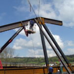 Ein Bauarbeiter steht auf einer Hubarbeitsbühne und arbeitet an einer großen Stahlkonstruktion, die im Freien zusammengebaut wird. Zwei weitere Arbeiter stehen darunter, im Hintergrund ist eine ländliche Landschaft mit blauem, bewölktem Himmel zu sehen. Kräne und Baumaterialien sind ebenfalls zu sehen.