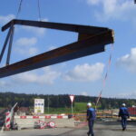 Auf einer Baustelle wird ein großer Stahlträger von einem Kran angehoben. Arbeiter in Schutzhelmen und Sicherheitsausrüstung stehen auf der Baustelle, einige gehen zu Fuß, andere beobachten. Warnschilder und Absperrungen sind sichtbar, und im Hintergrund sind Bäume und bewölkter Himmel zu sehen.