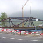 Eine Baustelle mit einem großen Stahlbrückengerüst im Bau. Hinter der Brücke steht ein Kran, und rot-weiße Absperrungen sperren den Bereich ab. Im Hintergrund ist ein nebliger Tag mit Hügeln und Bäumen zu sehen.