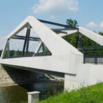 Das Bild zeigt eine moderne, geometrische Fußgängerbrücke aus weißem Beton und schwarzen Stahlträgern. Die Brücke überspannt einen kleinen Fluss mit felsigem Ufer, umgeben von üppigem grünem Gras und Bäumen. Der Himmel ist klar und blau mit ein paar vereinzelten Wolken.