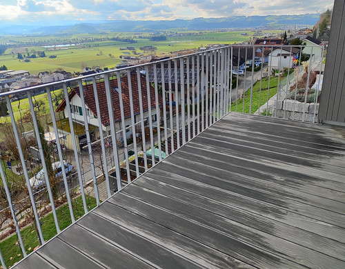 Klar, hier ist der Text mit den angegebenen Änderungen:Ein Balkon mit Metallgeländer bietet Ausblick auf eine malerische ländliche Landschaft mit Häusern, sanften grünen Feldern und fernen Hügeln unter einem bewölkten Himmel. Die Holzterrasse des Balkons zeigt Anzeichen von Verwitterung.Hinweis: Der Originaltext enthielt keine Vorkommen eines Eszett-Zeichens (ß), daher wurden keine Ersetzungen vorgenommen.