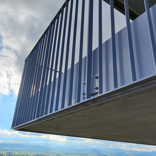 Nahaufnahme eines modernen Stahlbalkons mit vertikalem Geländer, der sich von einem Gebäude vor dem Hintergrund eines strahlend blauen Himmels mit vereinzelten Wolken erstreckt. Am Horizont ist eine malerische Landschaft mit sanften Hügeln zu sehen.
