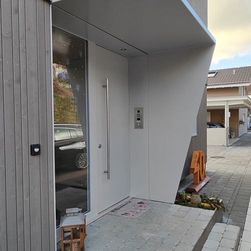Ein moderner Hauseingang mit einer grauen Tür mit vertikalem Griff. Die Tür ist von hohen Fenstern flankiert und hat eine kleine Markise darüber. Der Weg ist gepflastert und neben dem Eingang steht ein Holzhocker. Das Nachbarhaus und geparkte Autos sind sichtbar.