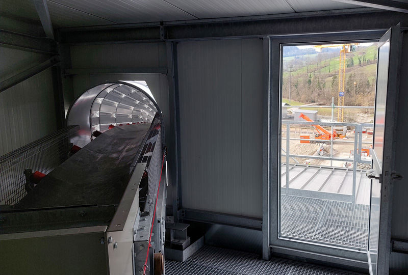 Ein Industrieraum mit Maschinen, darunter ein Förderband auf der linken Seite und eine große offene Tür auf der rechten Seite, die zu einem Außenbaubereich mit Kränen und Geräten führt. Die Außenszene zeigt im Hintergrund eine hügelige Landschaft.