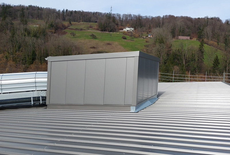 Auf einem Wellblechdach thront eine Metallkonstruktion, die von einem Sicherheitsgeländer umgeben ist. Im Hintergrund ist eine Hügellandschaft mit spärlichem Baumbestand und einigen Häusern unter einem teilweise bewölkten Himmel zu erkennen.