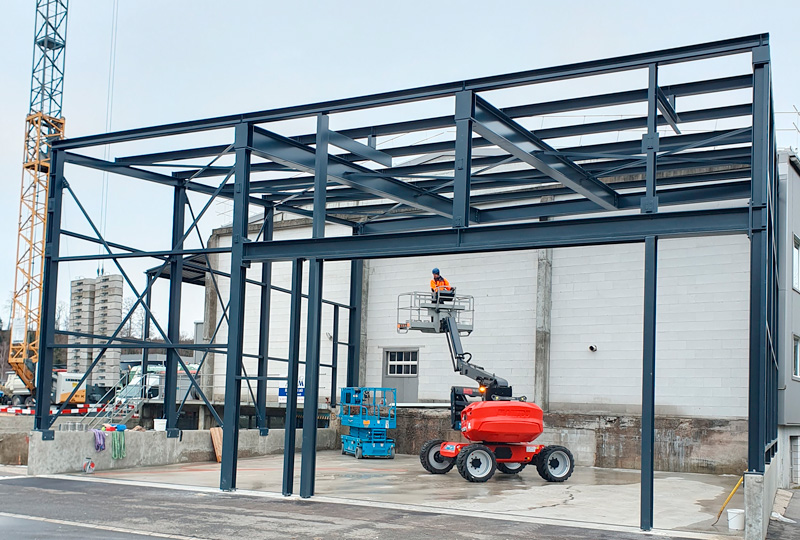 Baustelle mit einer unvollständigen Stahlrahmenkonstruktion. Ein Arbeiter in einer Warnweste bedient eine mobile Arbeitsbühne (MEWP), um auf den oberen Teil der Konstruktion zu gelangen. Auf der Baustelle sind Baumaterialien und Geräte zu sehen.