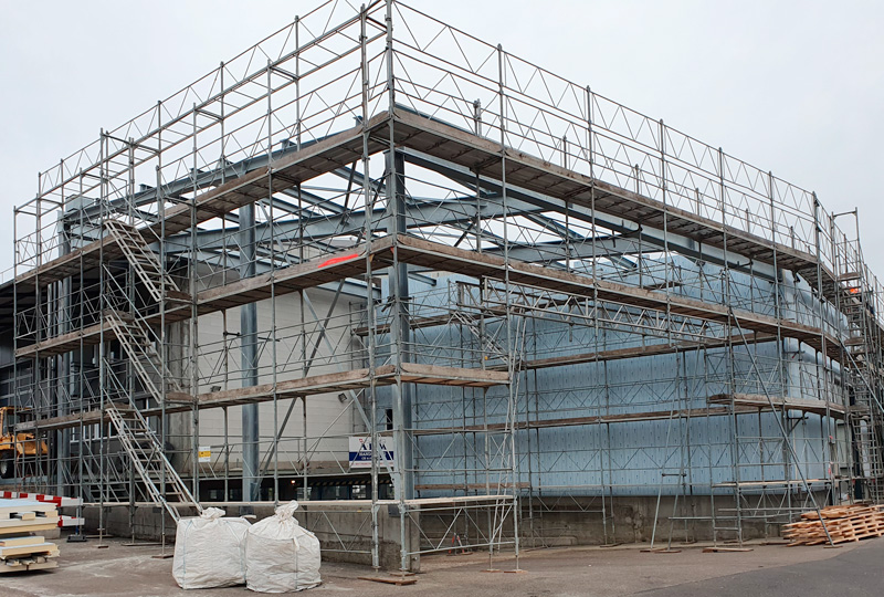 Eine Baustelle mit Gerüsten, die ein teilweise fertiggestelltes Gebäude umgeben. Das Gebäude scheint sich noch in der frühen Bauphase zu befinden, da Metallgerüste und provisorische Plattformen vorhanden sind. Auf der Baustelle sind verschiedene Baumaterialien und Geräte zu sehen.