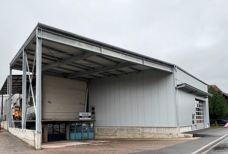 Ein großes Industrielager mit einer grauen Metallfassade und einem breiten Überhang am Eingang. Am Eingang hängt ein Schild mit der blauen Aufschrift „NETAL“. Der Bereich um das Lager umfasst eine gepflasterte Auffahrt und einen Blick auf einige Industrieanlagen im Inneren.