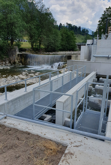 Ein Industriegebäude mit Metallstegen und Geländern liegt neben einem fließenden Fluss und einem kleinen Wasserfall in einem Waldgebiet mit Hügeln im Hintergrund. Der Himmel ist teilweise bewölkt, was die ruhige Landschaft noch verstärkt.