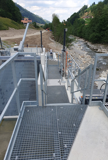 Ein Blick auf einen metallischen Gehweg und eine Struktur in der Nähe eines felsigen Flusses vor einem Hintergrund aus grünen Hügeln und Bäumen. Der Gehweg ist erhöht und hat Geländer auf beiden Seiten. Der Himmel darüber ist teilweise bewölkt und in der Ferne ist eine Baustelle sichtbar.