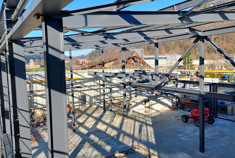 Eine Baustelle mit einem Stahlgerüst für ein großes Gebäude. Das Gerüst besteht aus mehreren Balken und Säulen. Im Hintergrund sind Kräne, Baumaschinen und Gebäude zu sehen. Die Baustelle ist vor einer Kulisse aus Bäumen und einem klaren blauen Himmel zu sehen.