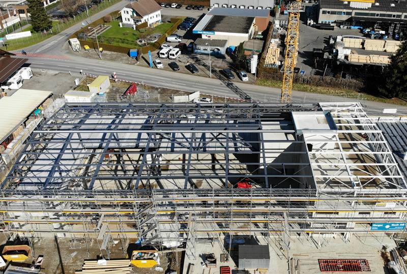 Luftaufnahme einer Baustelle mit einem Metallgerüst eines großen Gebäudes im Bau. Auf der Baustelle sind mehrere Baufahrzeuge und Materialien zu sehen. Das Gebiet umfasst nahe gelegene Gebäude, Straßen und einen Kran zum Heben schwerer Materialien.