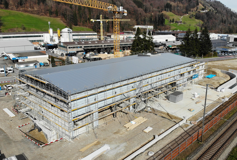 Ein großes Industriegebäude im Bau, umgeben von Gerüsten, daneben ein großer gelber Kran. Auf der Baustelle sind Baumaterialien und -geräte zu sehen. Im Hintergrund sind weitere Industriegebäude, eine Eisenbahnstrecke und sanfte grüne Hügel zu sehen.