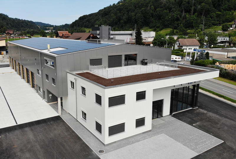 Eine Luftaufnahme eines modernen Industriegebäudes mit Flachdach. Das Gebäude hat eine Kombination aus grauen und weißen Außenpaneelen und verfügt über Solarmodule auf dem Dach. Um das Gebäude herum befinden sich weitere Industrie- und Wohngebäude, vor der Kulisse grüner Hügel.