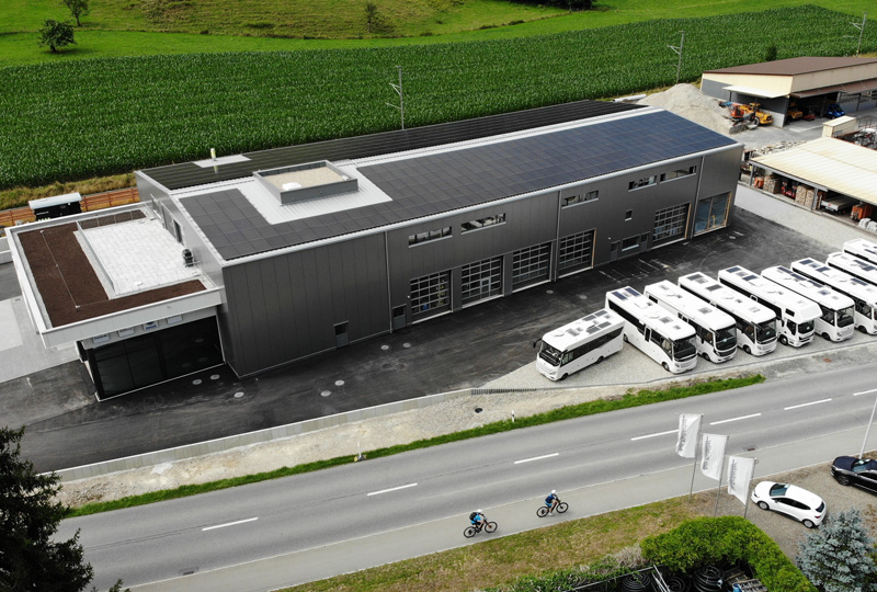 Eine Luftaufnahme eines modernen Lagerhauses mit einem Solarpanelendach, mehreren daneben geparkten weißen Bussen und einer Straße mit zwei Radfahrern im Vordergrund. Grüne Felder und verschiedene Nebengebäude umgeben das Lagerhaus.