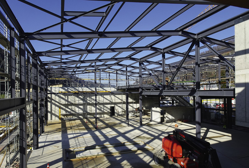 Ein teilweise fertiggestelltes Gebäude mit Stahlgerüst unter einem klaren blauen Himmel. Schatten der Struktur werden auf den Betonboden geworfen. Auf der linken Seite ist ein Gerüst zu sehen und im Vordergrund rechts ist ein rotes Baufahrzeug geparkt.