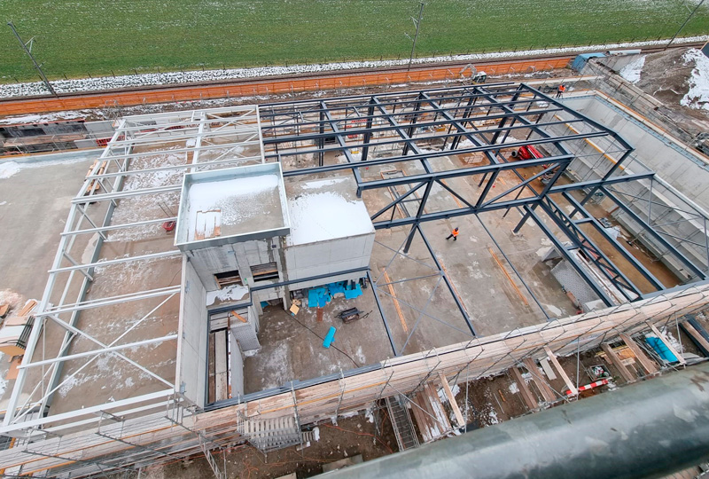 Ihr Text enthält keine Vorkommen des Eszett-Zeichens (ß), daher sind keine Ersetzungen erforderlich. Hier ist der Originaltext zu Ihrer Information:„Luftaufnahme einer Baustelle mit einer Stahlrahmenkonstruktion. Der Bereich hat teilweise fertiggestellte Böden, einige Baumaterialien sind verstreut und auf Teilen der Baustelle liegt Schnee. Arbeiter und Ausrüstung sind zu sehen und im Hintergrund befindet sich ein grünes Feld.