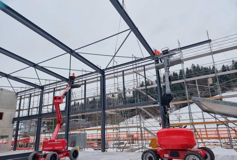 Bauarbeiter montieren mit roten und gelben Hubarbeitsbühnen Stahlträger für das Gerüst eines Gebäudes. Die Baustelle ist von Gerüsten und schneebedecktem Boden umgeben, im Hintergrund sind bewaldete Hügel unter einem wolkigen Himmel zu sehen.