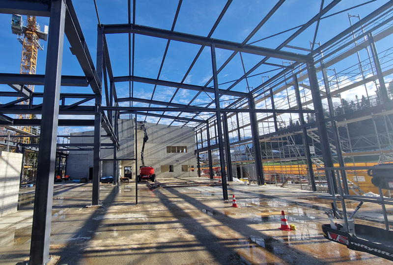 Eine Baustelle mit dem Stahlgerüst eines großen Gebäudes unter einem klaren blauen Himmel. Verschiedene Baugeräte und Materialien liegen verstreut herum, im Hintergrund sind Gerüste zu sehen. Auf dem nassen Boden stehen orangefarbene Leitkegel.