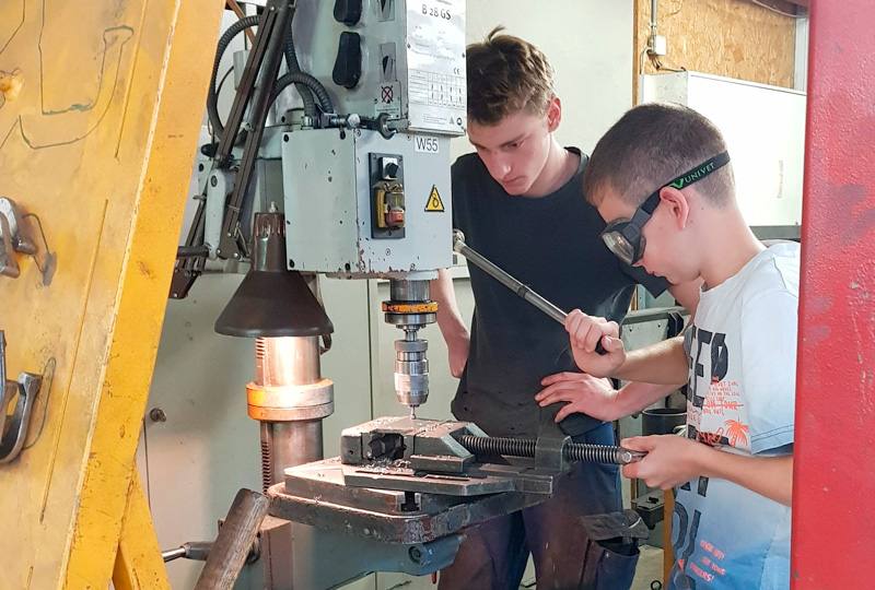 Zwei junge Menschen mit Schutzbrillen bedienen eine Metallbearbeitungsmaschine in einer Industriewerkstatt. Einer bohrt ein an der Maschine befestigtes Stück Metall, während der andere beim Einstellen hilft. Im Hintergrund sind Maschinen und Werkstattwerkzeuge zu sehen.
