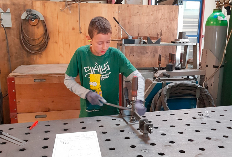 Ein kleiner Junge, der ein grünes T-Shirt mit einer Comicfigur und Handschuhe trägt, arbeitet in einer Werkstatt. Er benutzt Werkzeuge auf einer Metallwerkbank, auf der überall Pläne und andere Materialien verstreut sind. Im Hintergrund sind verschiedene Geräte und Holzoberflächen zu sehen.