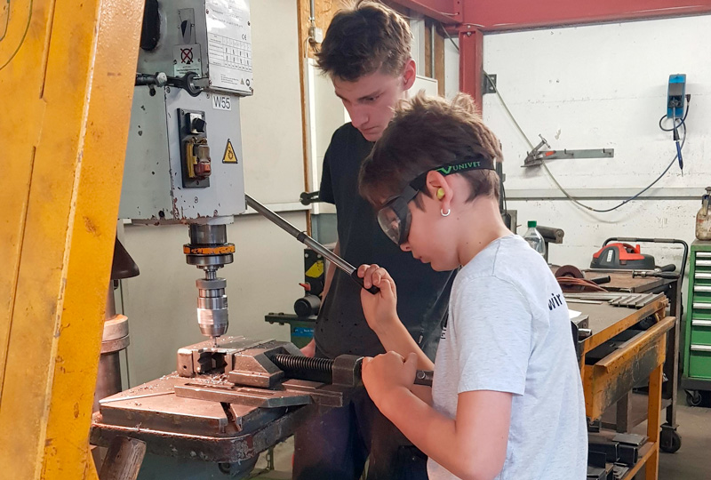 Zwei Personen arbeiten in einer Werkstatt an einer Metallbohrmaschine. Die jüngere Person mit Schutzbrille bedient die Maschine, während die ältere Person aufmerksam zusieht und Anleitung gibt. Im Hintergrund sind verschiedene Werkzeuge und Werkstattausrüstung zu sehen.