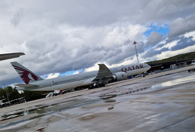 Ein Flugzeug von Qatar Airways steht auf einer nassen Landebahn eines Flughafens. Der Himmel ist bedeckt, zwischen den Wolken sind blaue Flecken zu sehen. Auf der nassen Oberfläche sind Spiegelungen des Flugzeugs und des Himmels zu sehen. Im Hintergrund sind verschiedene Flughafenfahrzeuge und -geräte zu sehen.