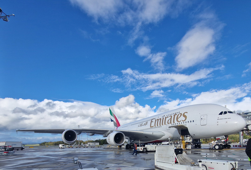 Ein Airbus A380 von Emirates steht auf dem Rollfeld unter einem strahlend blauen Himmel mit vereinzelten Wolken. Das Logo der Fluggesellschaft ist an der Seite des Flugzeugs zu sehen. Fahrzeuge und Ausrüstung des Bodendienstes sind rund um das Flugzeug verteilt.