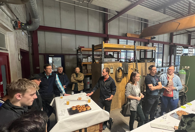 Eine Gruppe von Menschen steht um einen Tisch mit Essen und Getränken in einer lagerhallenähnlichen Umgebung. Der Raum ist mit Industrieregalen und -geräten ausgestattet, mit roten Balken und weißen Wänden. Einige Teilnehmer unterhalten sich, während andere aufmerksam zuhören.
