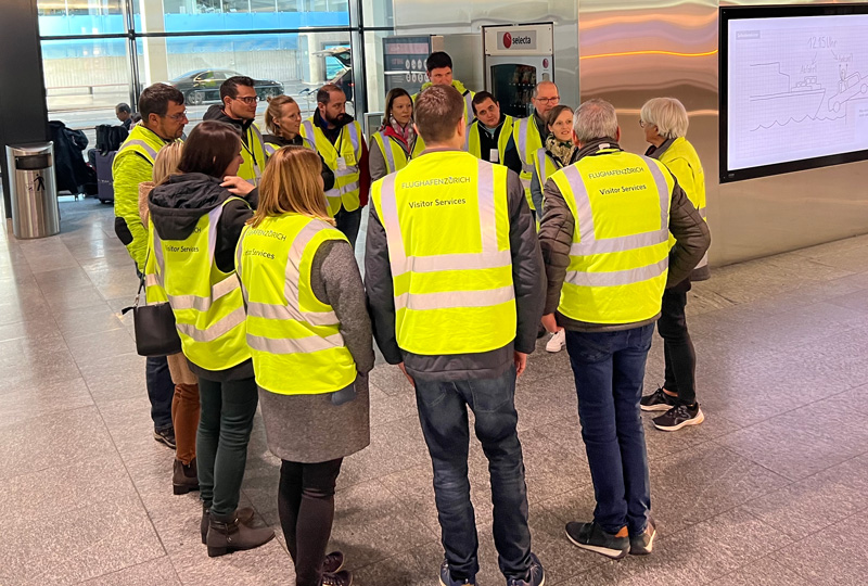 Eine Gruppe von Menschen in leuchtend gelben „Visitor Services“-Sicherheitswesten steht in einem Kreis in einem Innenbereich. Sie scheinen in eine Diskussion vertieft zu sein oder Anweisungen entgegenzunehmen. An der Wand rechts ist eine große Karte oder ein Diagramm zu sehen.