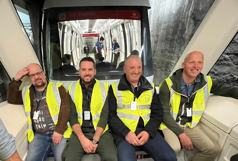 Vier Männer in Warnwesten sitzen lächelnd in einem Flughafen-Transitfahrzeug. Sie scheinen Teil einer Führung oder Inspektionsgruppe zu sein. Hinter ihnen ist durch eine Glasscheibe das Innere eines anderen Fahrzeugs und im Hintergrund ein Tunnel zu sehen.