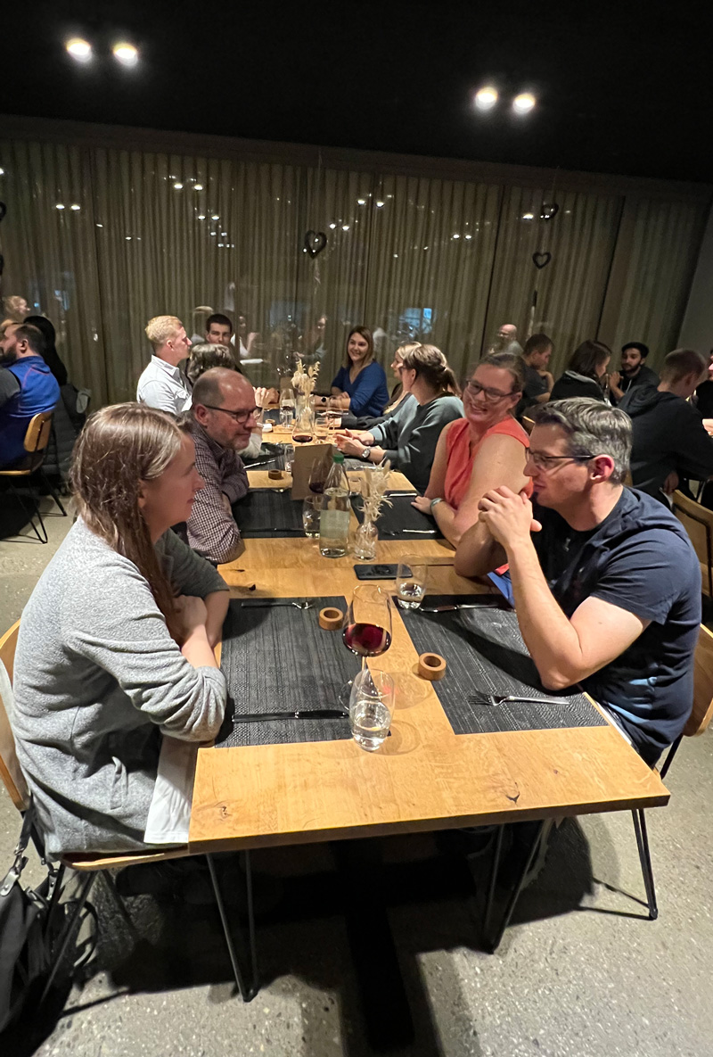 Eine Gruppe von Menschen sitzt an einem langen Holztisch in einem schwach beleuchteten Restaurant. Sie unterhalten sich, lächeln und genießen ihre Zeit. Im Vordergrund unterhalten sich zwei Paare über den Tisch hinweg. In der Mitte steht ein Glas Rotwein.