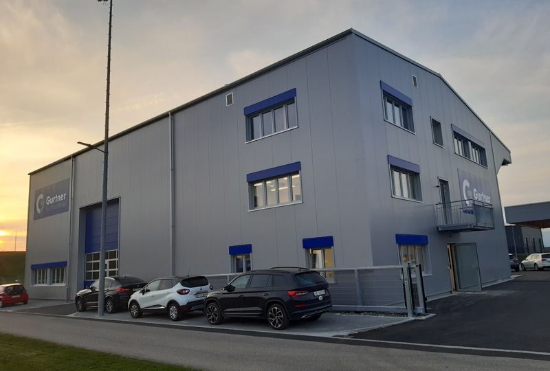 Ein modernes, graues Geschäftsgebäude mit mehreren Fenstern. Drei Autos sind vor dem Gebäude geparkt. Die Fenster sind mit blauen Akzenten versehen und in der Nähe des Eingangs befindet sich ein Logo. Neben dem Gebäude ist ein hoher Mast zu sehen und der Himmel scheint bei Sonnenuntergang bewölkt zu sein.