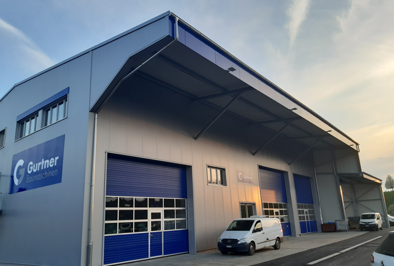 Ein großes Industriegebäude mit dem Namen „Guertner“ und dem Logo an der Fassade. Das Gebäude ist in Blau und Grau gehalten, hat mehrere Garagentore und davor parkt ein weißer Lieferwagen. Es ist Tag und der Himmel ist teilweise bewölkt.