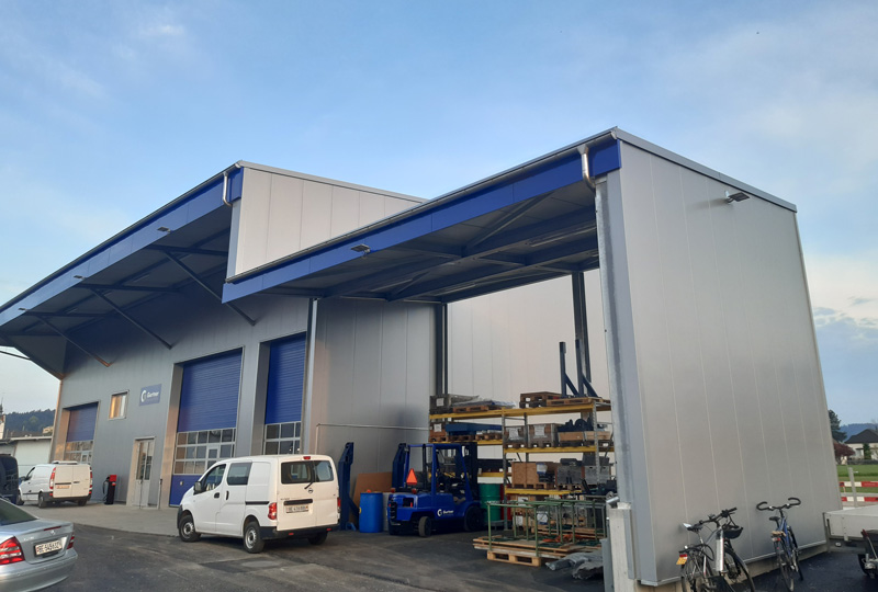 Eine große Industriegarage mit offenen blauen Toren. Mehrere Fahrzeuge, darunter ein weißer Lieferwagen und Pkw, sind draußen geparkt. Verschiedene Maschinen und Geräte sind unter dem offenen Unterstand neben dem Hauptgebäude gelagert. In der Nähe sind auch Fahrräder zu sehen.