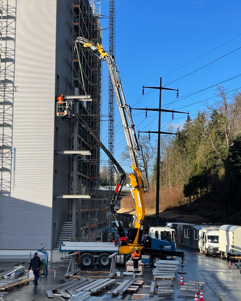 Eine Baustelle mit einem großen gelben Kran, der Arbeiter in einem Korb an die Fassade eines mehrstöckigen Gebäudes hebt. Das Gebäude ist teilweise mit Gerüsten bedeckt. Arbeiter und Fahrzeuge sind da und der Boden ist mit Baumaterialien verstreut. Im Hintergrund sind Bäume zu sehen.