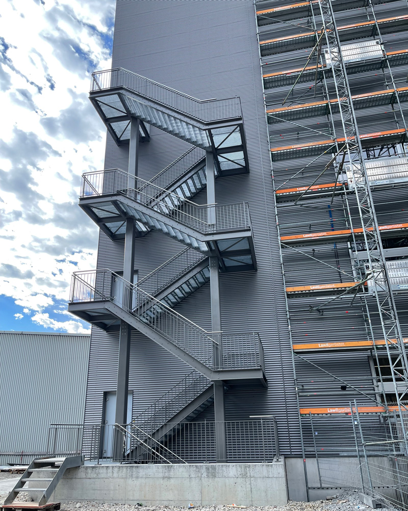 Ein hohes, graues Gebäude mit einer außenliegenden Feuertreppe aus Metall. Die Treppe verläuft im Zickzack an der Seite des Gebäudes entlang nach oben. Der Himmel darüber ist teilweise bewölkt und auf der rechten Seite des Gebäudes ist ein Gerüst zu sehen.