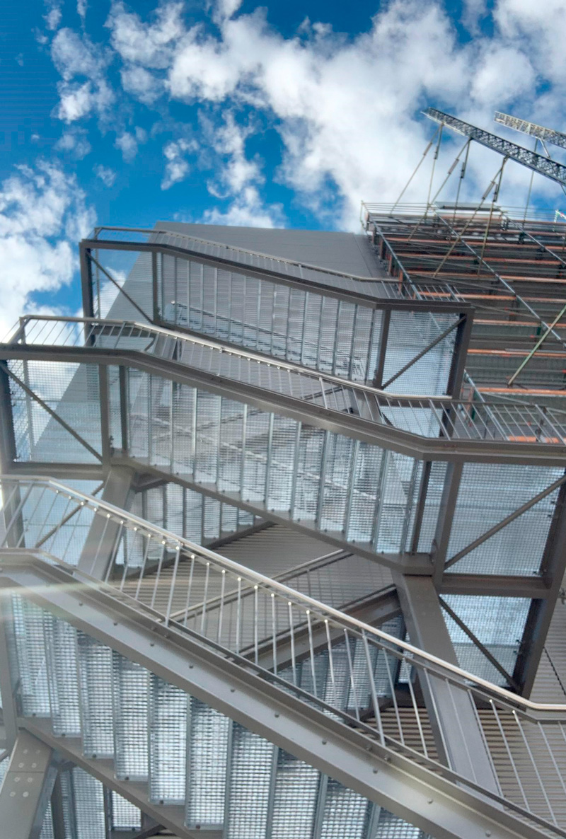 Ein Foto einer Metalltreppe im Freien an einem modernen Gebäude, die über mehrere Zickzack-Flüge nach oben führt. Das Gebäude ist in einen hellen, klaren Tag eingebettet, über dem sich vereinzelte Wolken am blauen Himmel befinden, was ein Gefühl von Offenheit und modernem Architekturstil vermittelt.