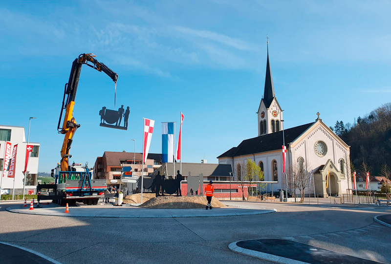In Ihrem Text kommt das Eszett-Zeichen nicht vor, daher sind keine Ersetzungen erforderlich. Hier ist der Text in seiner jetzigen Form:Ein großer Kran installiert eine Metallskulptur an einem Kreisverkehr vor einer Kirche mit einem hohen Kirchturm. Ein Arbeiter in einer orangefarbenen Weste steht in der Nähe, während im Hintergrund einige Gebäude und Fahnenmasten mit rot-weißen Flaggen zu sehen sind. Der Himmel ist klar und blau.