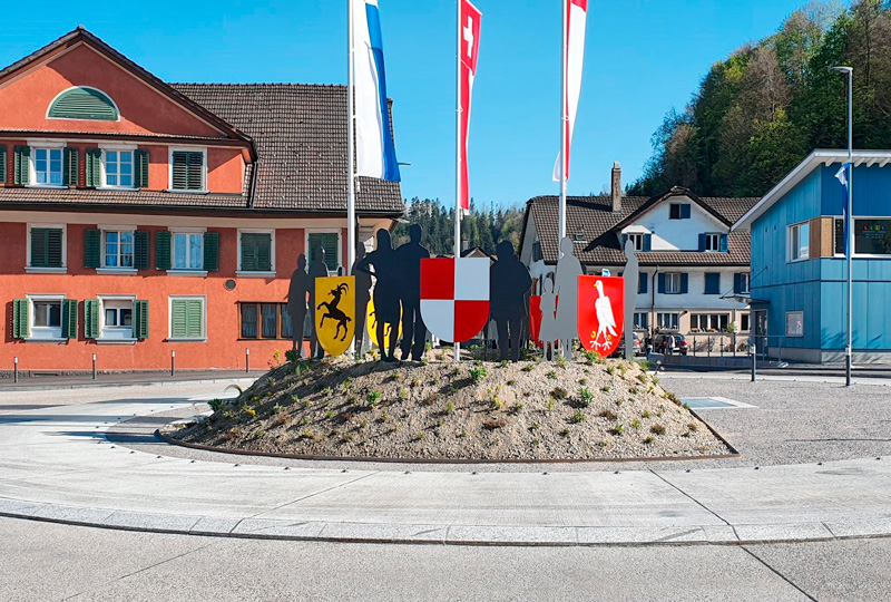Vor einer bunten Kulisse aus Wohnhäusern steht ein Kreisverkehr mit metallischen Menschensilhouetten und verschiedenen regionalen Emblemen. Im Vordergrund sind ein rotes Gebäude mit grünen Fensterläden und Blumenkästen und ein blaues Gebäude mit großen Fenstern zu sehen. Im Hintergrund sind grüne Hügel zu sehen.