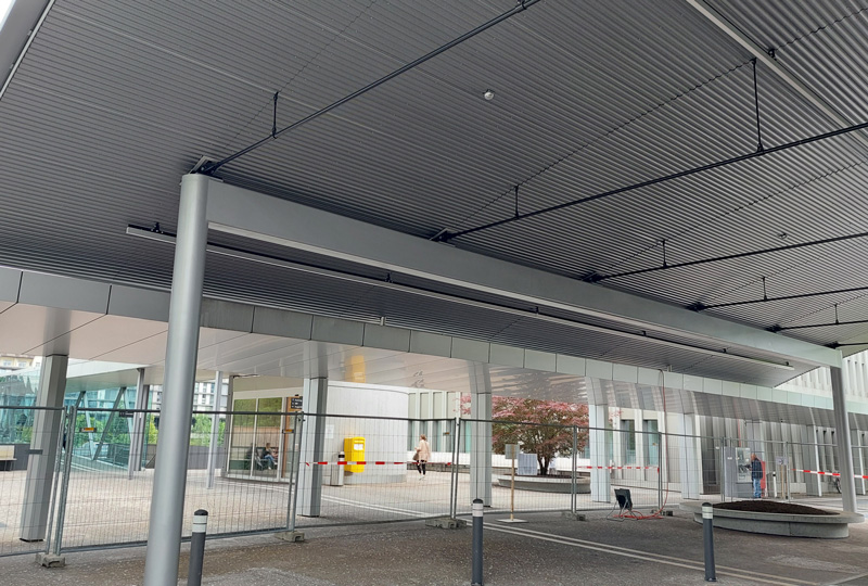 Ein überdachter Durchgang mit Metallträgern und einem grauen Wellblechdach, mit einem eingezäunten Bereich. Im Hintergrund sind Gebäude und ein kleiner Baum zu sehen. In der Ferne sieht man ein paar Leute gehen.