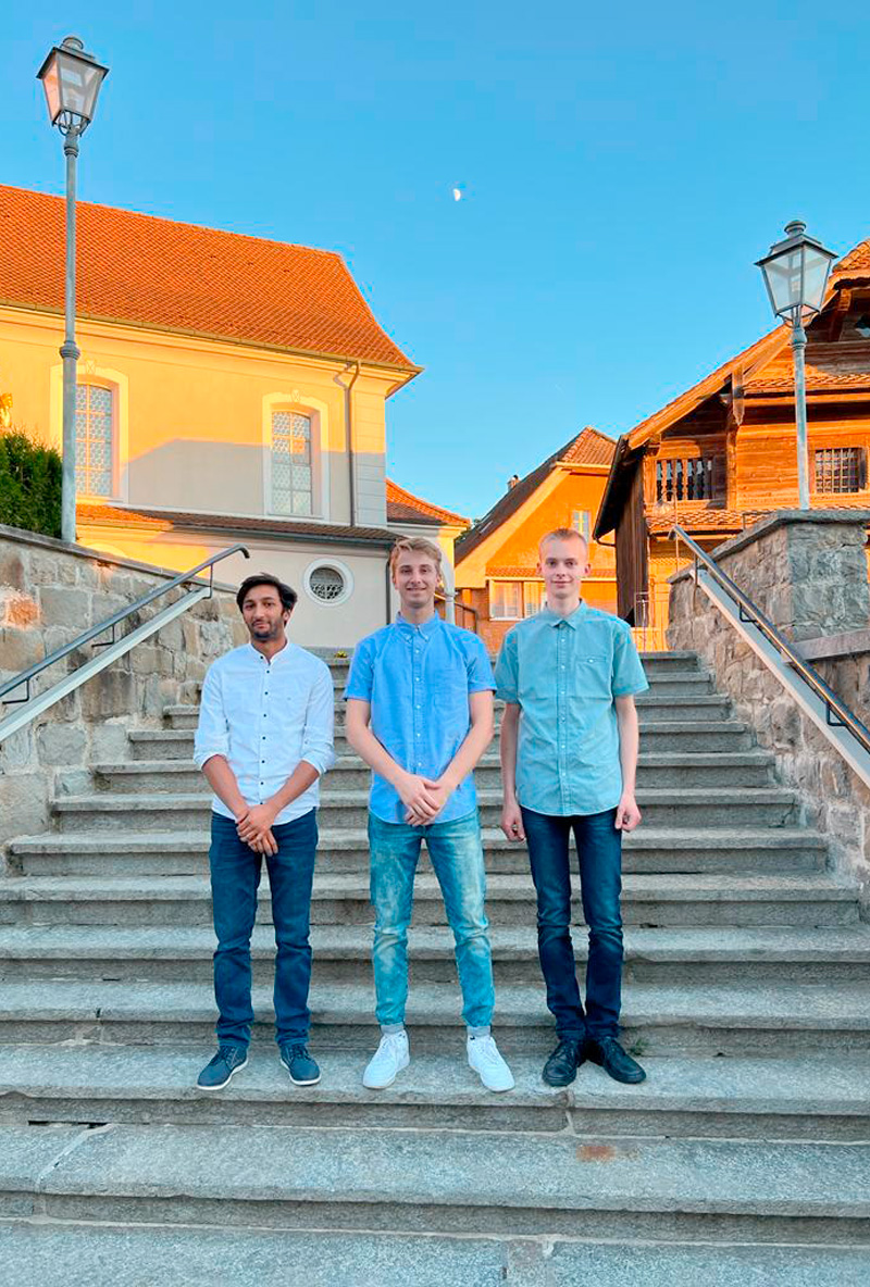 Drei junge Männer stehen auf einer breiten Steintreppe im Freien. Im Hintergrund sind Häuser und Straßenlaternen zu sehen. Sie tragen alle legere Hemden und Jeans. Der Himmel ist klar und blau, was darauf schließen lässt, dass es ein sonniger Tag ist.