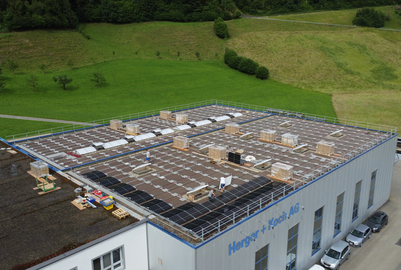 Luftaufnahme eines großen Gebäudes mit mehreren auf dem Flachdach installierten Solarmodulen. Mehrere Arbeiter sind anwesend und verschiedene Geräte sind überall verstreut. Im Hintergrund sind grüne Hügel und Grasflächen zu sehen. Auf der Wand des Gebäudes steht der Text „Hengst + Kunz AG“.
